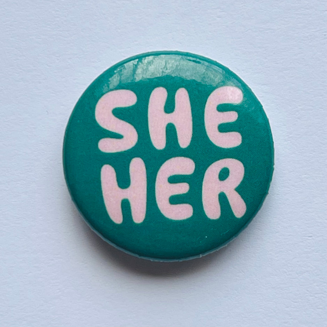 one inch lapel button reads "She Her" in funny blush pink lettering on emerald green